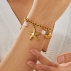 Gold-plated charm necklace worn on wrist: starfish pendant, shell charm, and white pearl beads