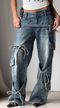 Blue denim utility jeans with long side straps, patch pockets, and knot ties at seams.