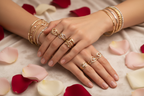 Hands wearing gold bangles and multiple rings, with flower petals around.