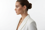 Gold-tone pendant necklace and dangling earrings on model side profile, white jacket.